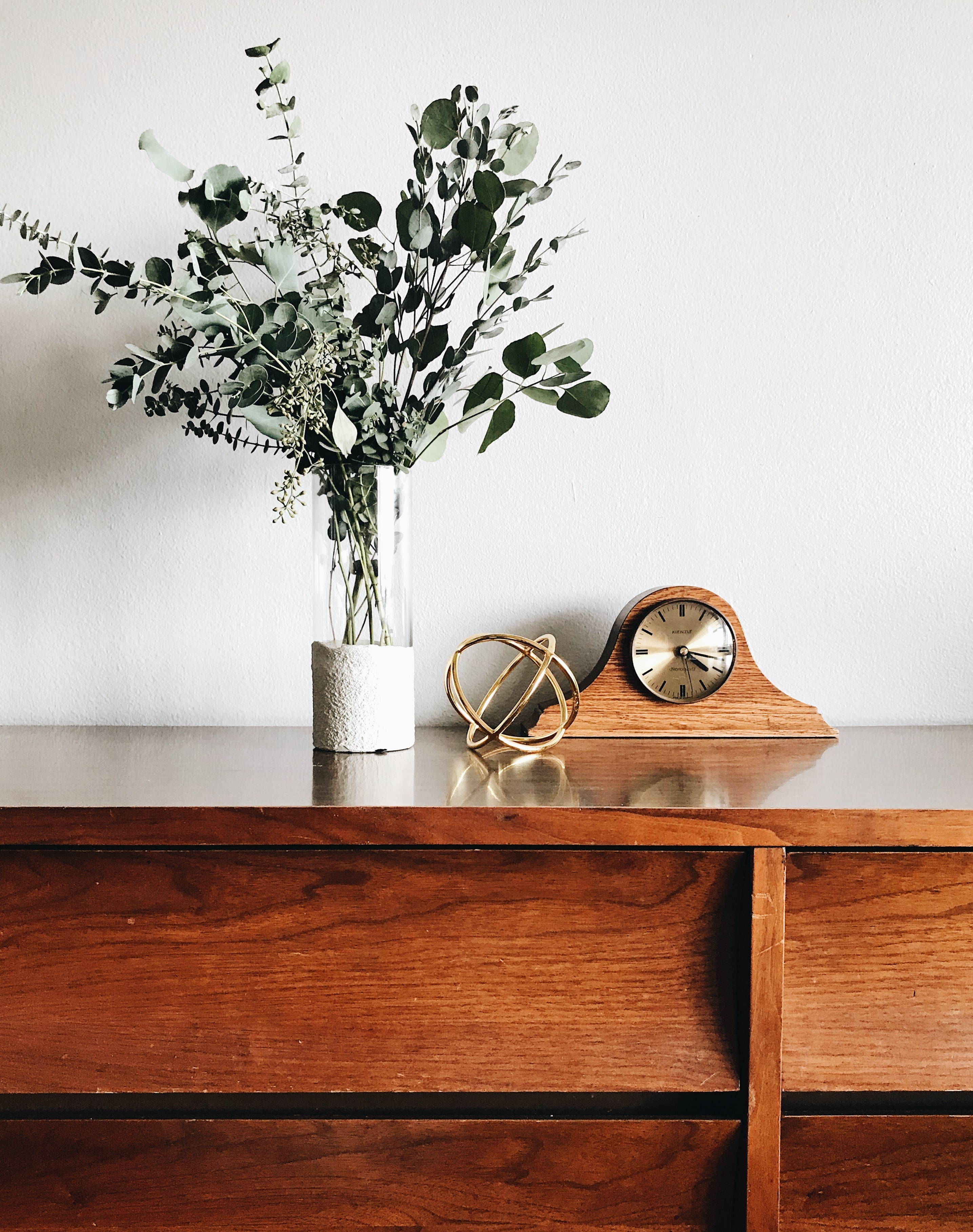 Light brown wooden dresser. There is a vase with greenery, a sculpture, and a vintage-looking table clock on the dresser.