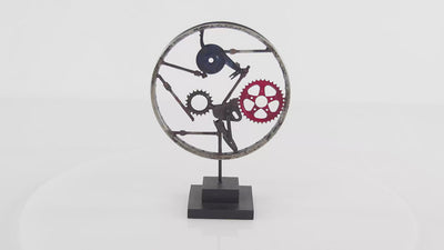 Sculpture made of solid metal and features a black wooden stand, rusty silver weal, and red and black gear-inspired details in the middle of a wheel. Sculpture is spinning 360 degrees and background is all white