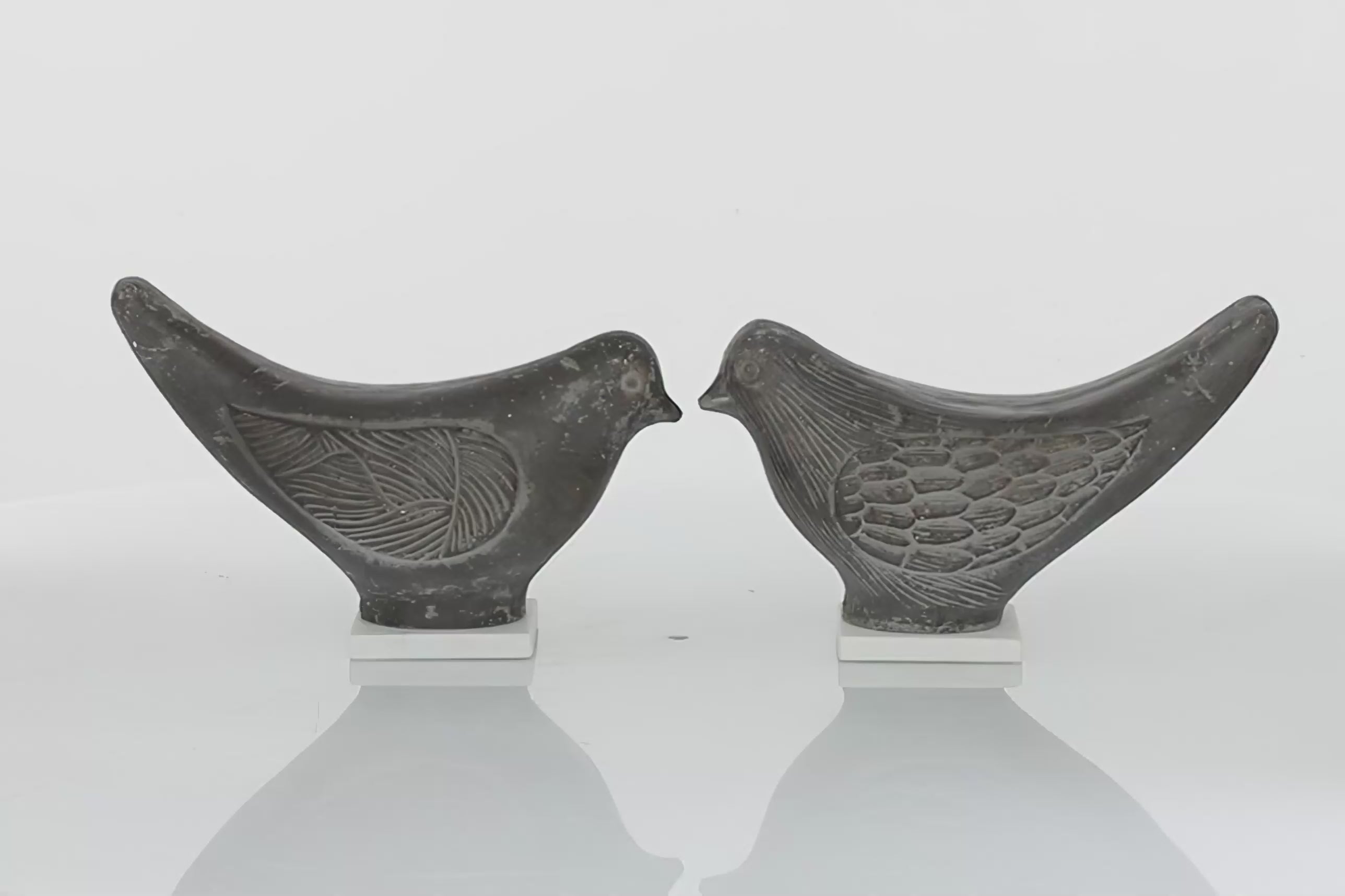 Set of two grey bird figurines textured on the face, wing and tail areas. Textures are in gold color. Birds are placed on the squared white marble stand. Birds are facing each other and spinning in 360 degrees. Background is all white
