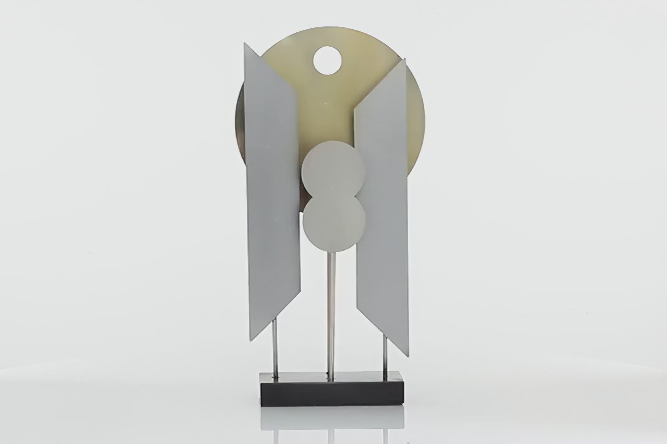 Grey and gold colored abstract aluminum figurine. Sculpture has a Gold circle and two grey parts that look wings. Figurine is spinning in 360 degrees and has a black wooden stand. Back ground is white.