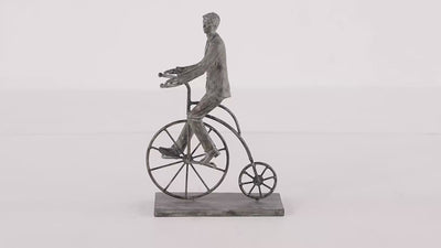 Sculpture that features a cyclist on a penny-farthing-inspired bicycle balanced securely on a flat platform base. Figurine is in grey color and is spinning 360 degrees. Background is all white