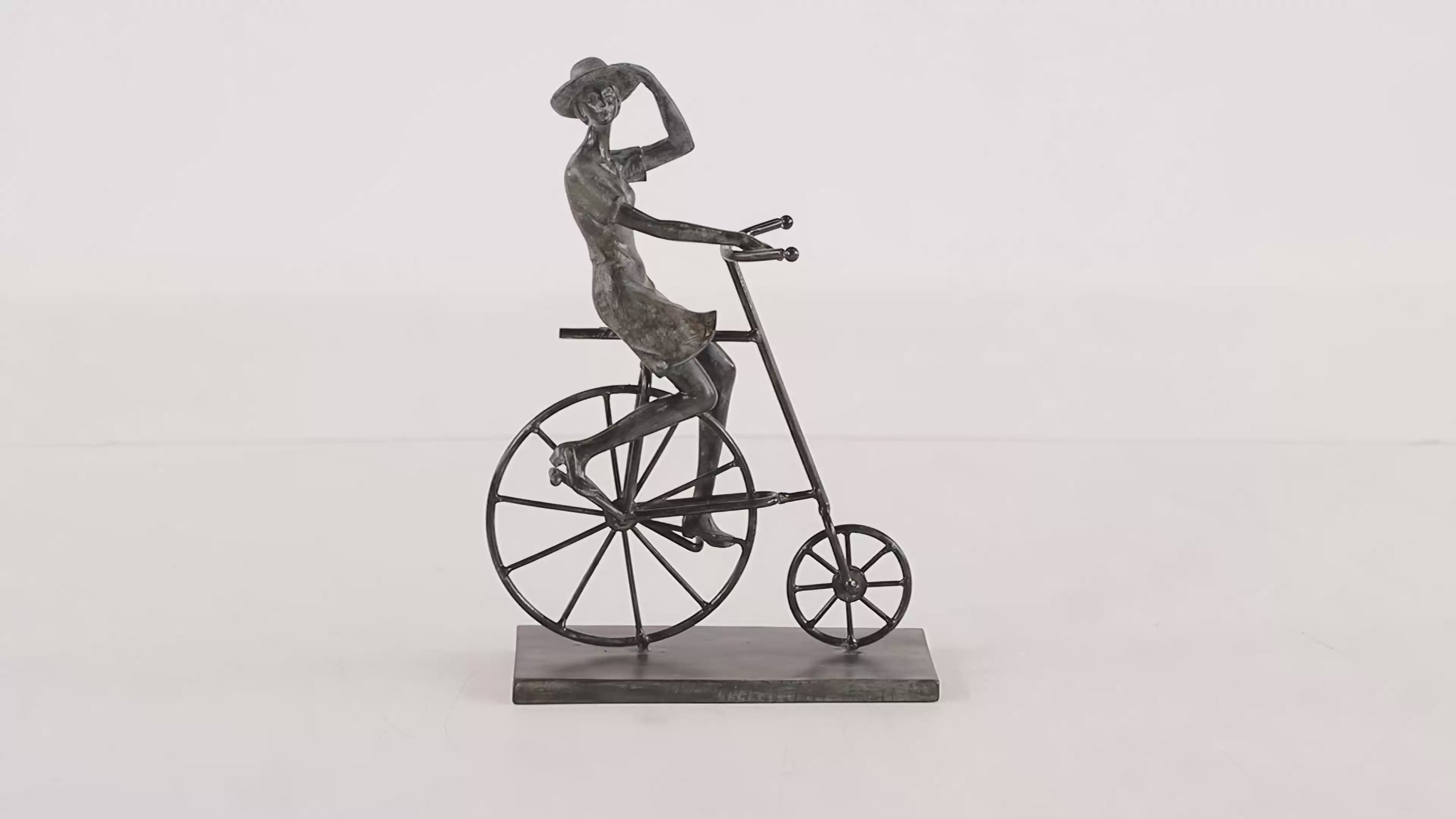 Sculpture that features a cyclist on a penny-farthing-inspired bicycle balanced securely on a flat platform base. Figurine is in grey color and is spinning in 360 degrees. Background is all white