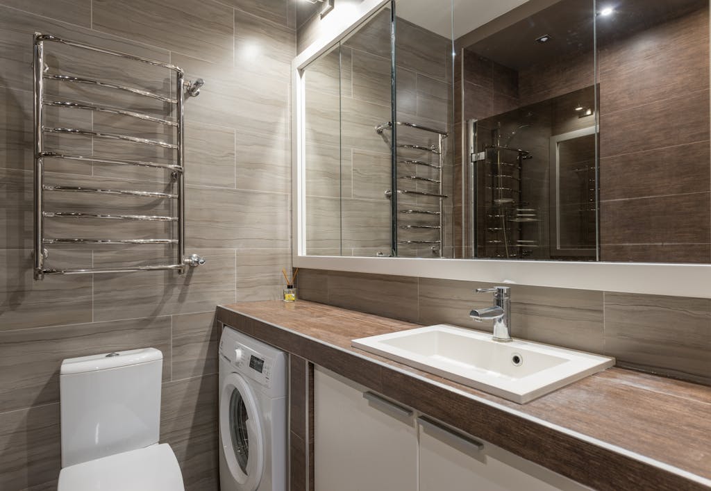 White ceramic toilet bowl and sink in contemporary bathroom with big mirror reflecting brown tiled walls