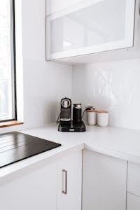 Modern electric coffee maker placed on white counter in kitchen near black stove and cupboards in light apartment with window